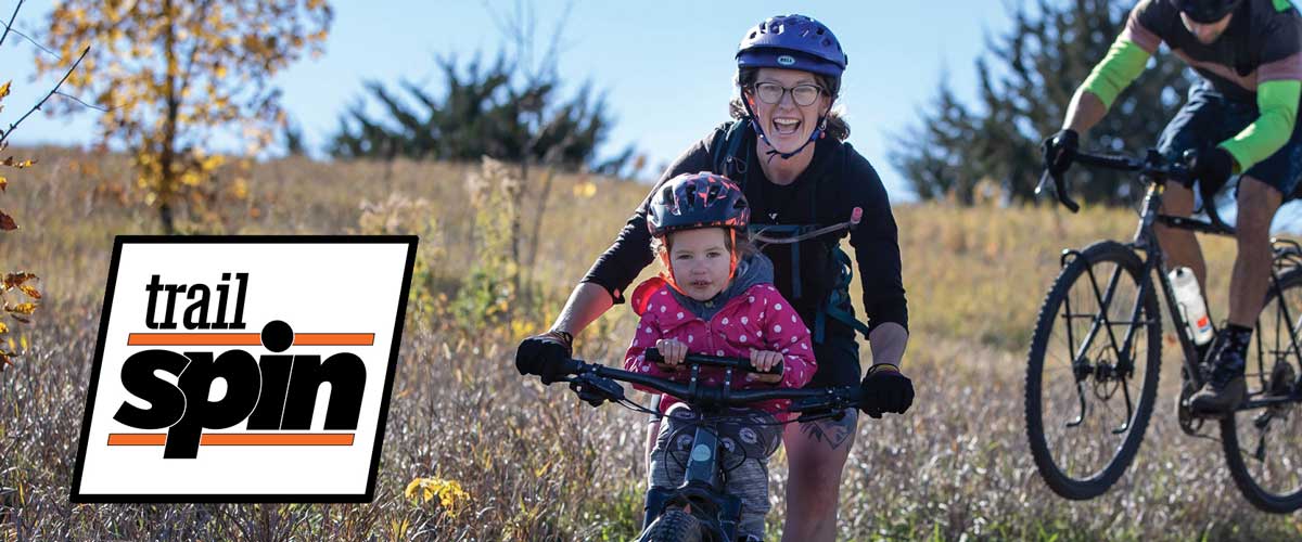 A mother and child smile while mountain biking. A Trail Spin logo is on the left side of the photo.