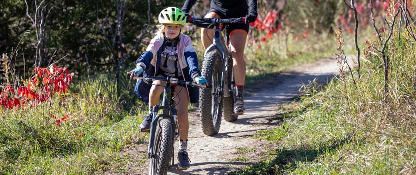 A kid rides a mountain bike down a trail with their parent behind them.