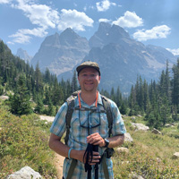 Tyler, wears a plaid shirt, hat and backpack and holds hiking poles while standing in front of mountains in the Teton Range.