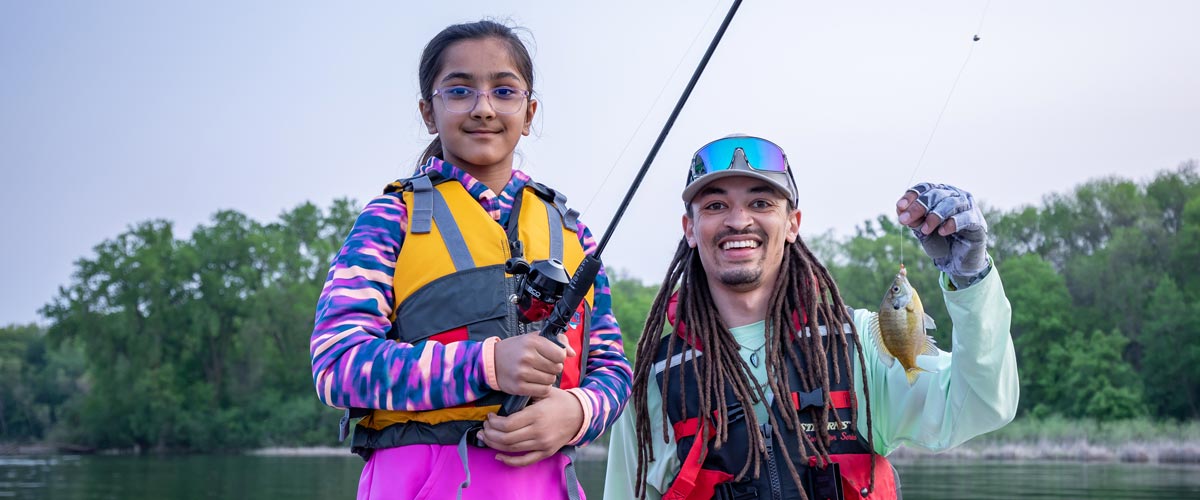 A girl holds a fishing pole. Next to her, a man smiles while holding the line of her fishing pole with a sunfish caught on the end.