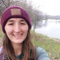 Zoe wears a burgundy, knit beanie over long brown hair and smiles, in front of a lake.