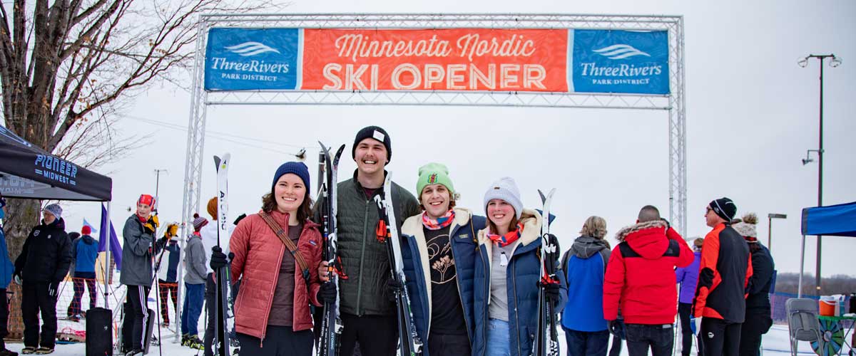 A group of people dressed in winter clothing hold skis in front of the Minnesota Nordic Ski Opener sign.