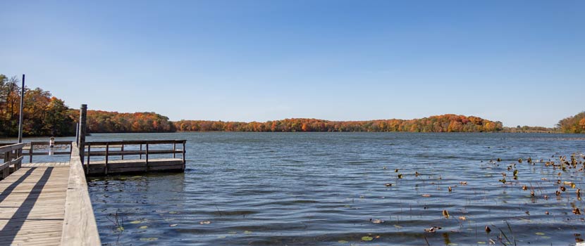 Photo of a pier on a lake. Shoreline trees' leaves are changing.