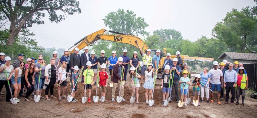 people stand with shovels in front of a large construction digger with trees in the background