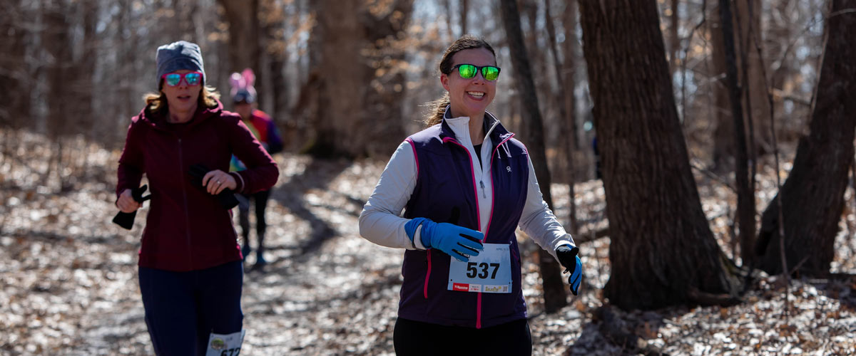 A runner wearing sunglasses smiles as she runs on a leaf-covered trail. Runners behind her.