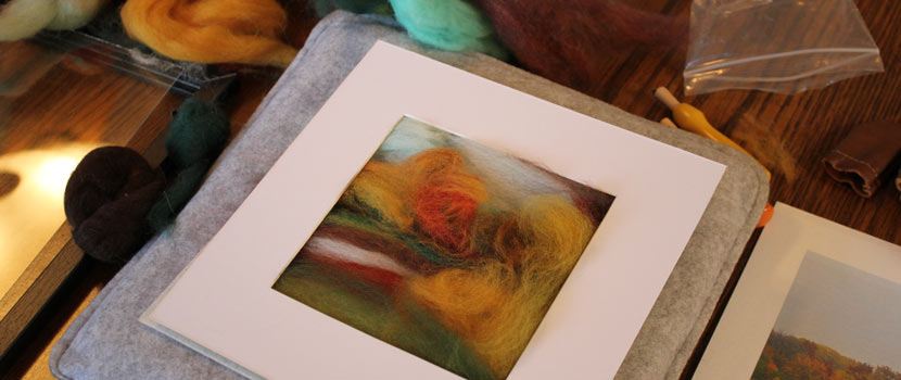 Photo of a felted landscape in progress. Roving is swirled inside of a frame, and dry felting tools and roving are laying nearby on the table.