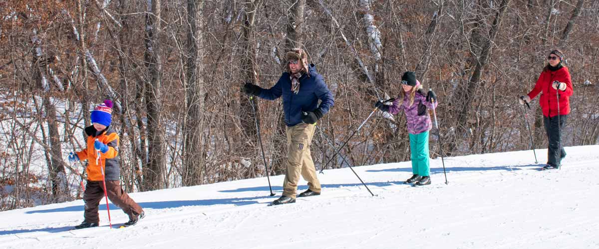 Photo of four people, a mix of adults and children, cross-country skiing on a trail with bare trees in the background.
