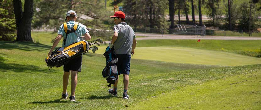 Two boys carry golf clubs across a golf course.