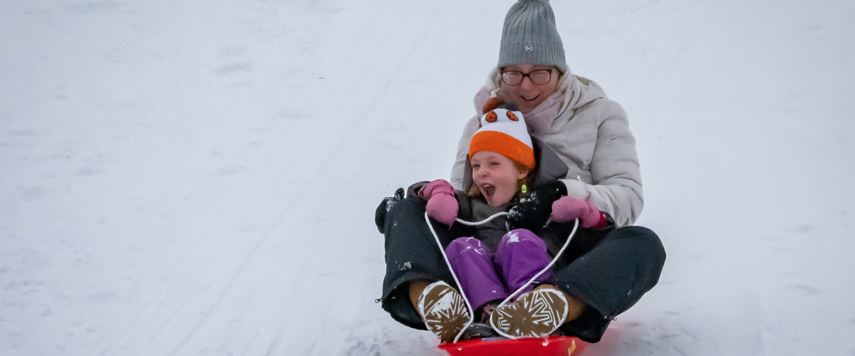 A photo of a mother and child sledding down a snowy hill; the child has a joyful face.