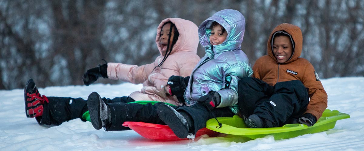 A group of three kids sit on plastic sleds at the top of a snowy hill.