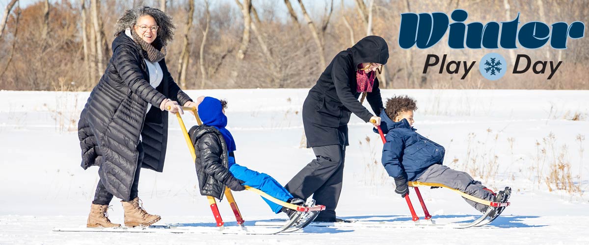 Photo of two adults pushing two children in kicksleds across snow with bare trees in the background. The Winter Play Day logo sits in the right upper corner of the image.