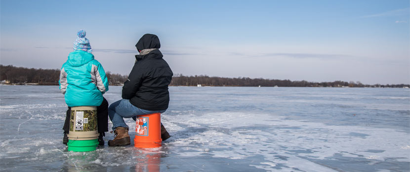 two people sitting on upside down 5-gallon pails on a frozen lake while ice fishing