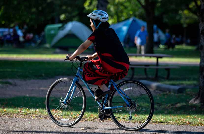 A girl wearing a bike helmet over a headscarf rides a blue bike down a paved path. A picnic table and campsite with tents are out-of-focus in the background.