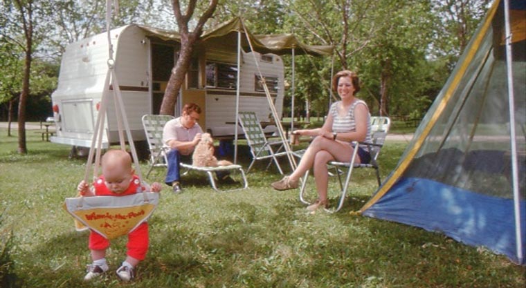 A man and woman sit at a picnic table in front of an RV while a baby plays in a swing in front of them. 