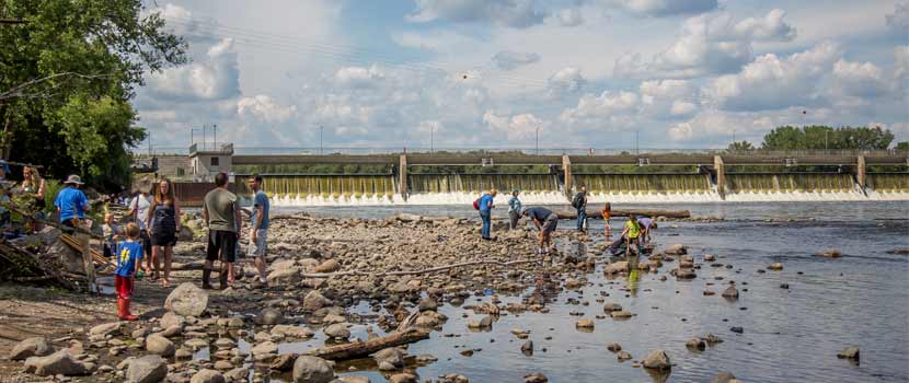People walk along the rocky shore of the Mississippi River. The Coon Rapids Dam can be seen behind them.