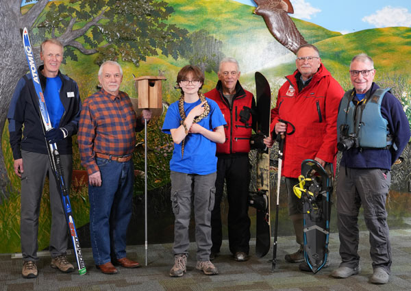 The 2024 Distinguished Volunteers stand with props related to their service, including skis, a snake, snowshoes and a camera.