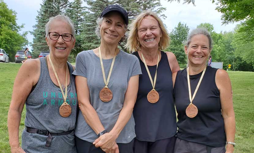 Leader Birdie, Co-founder Diane, Leader Bev and Co-founder Teresa celebrate one year of hiking in the Hyland Sole Mates Hiking Club on July 9, 2021. They wear wooden medallions with Hyland Sole Mates info etched on them.