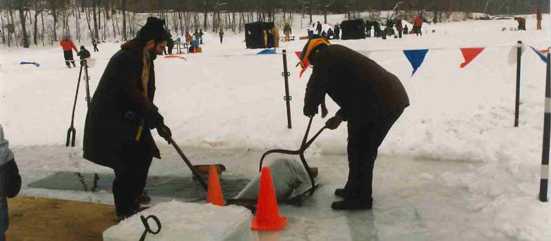 Two people use tools to lift cut ice out of the water. Behind them, people ice fish and play on the snow.