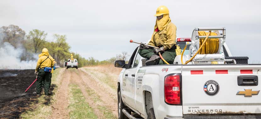 a person in a yellow hard hat and fire-resistant suit sits in the bed of a pick-up truck with a hose in hand. 