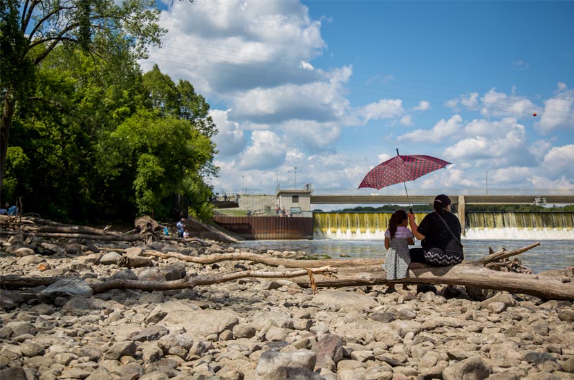 A woman and her daughter sit on a log on the banks of a MIssissippi River.