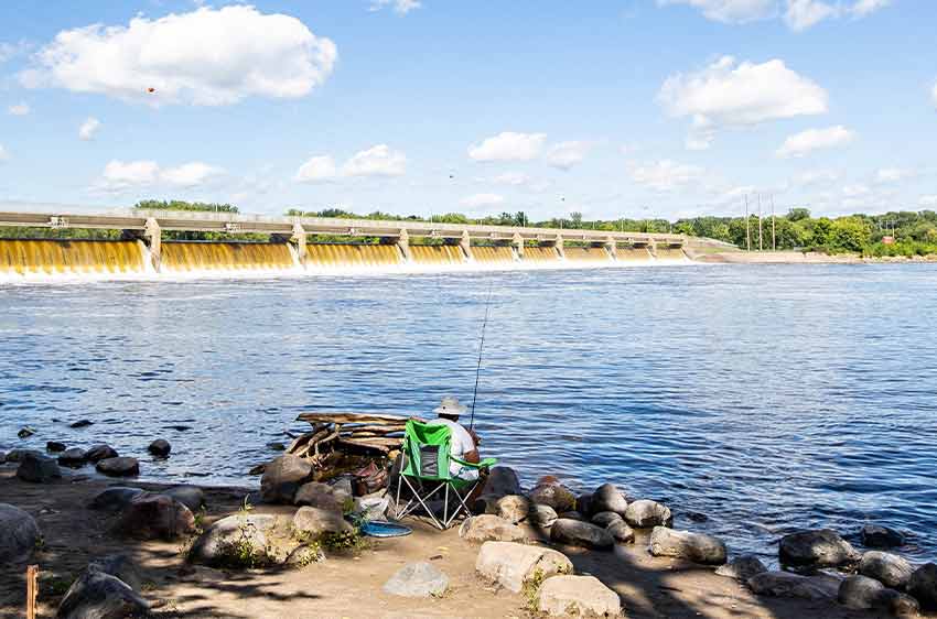 A person sits in a chair fishing on the rocky bank of the Mississippi River. A dam spanning the river is in the background.
